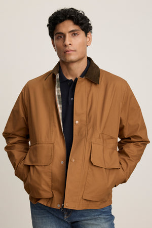 A man poses against a plain background in blue jeans and the DESREN JACKET by Velvet by Graham & Spencer, featuring brown cotton canvas, a corduroy collar, and large front pockets.