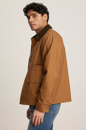 A person wearing the Velvet by Graham & Spencer DESREN JACKET, a brown cotton canvas style with a corduroy collar, and blue jeans stands against a plain light background, facing slightly to the side.