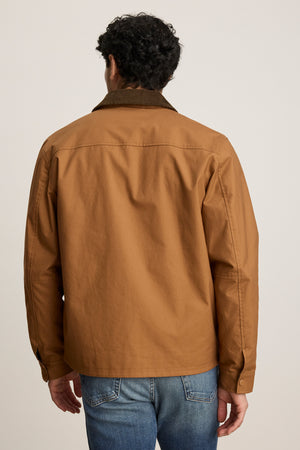 A person is shown from the back, standing against a plain light background, wearing the DESREN JACKET by Velvet by Graham & Spencer—a brown cotton canvas jacket with a corduroy collar—and blue jeans.