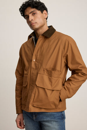A person wears the DESREN JACKET by Velvet by Graham & Spencer, a brown cotton canvas jacket with large front pockets and a corduroy collar, paired with blue jeans, standing against a plain background.