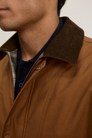 Close-up of a person wearing the DESREN JACKET by Velvet by Graham & Spencer—a brown cotton canvas jacket with a dark corduroy collar and plaid lining, layered over a dark shirt. The person's face is partially visible.