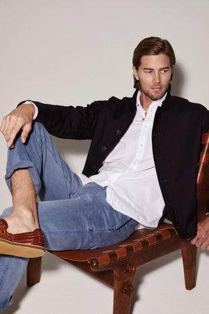 A man sitting on a wooden chair wearing TOBY SANDED TWILL PANT by Velvet by Graham & Spencer and a jacket.