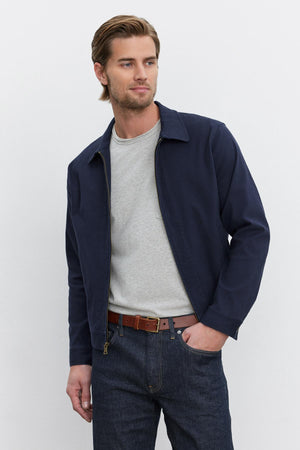 A man with brown hair wearing a navy STEPHAN JACKET by Velvet by Graham & Spencer over a grey shirt, blue jeans, and a brown belt, standing against a plain white background.
