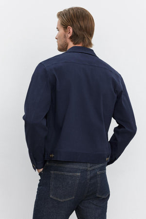 A person with light hair is standing with their back to the camera, wearing a dark blue STEPHAN JACKET by Velvet by Graham & Spencer and blue jeans.