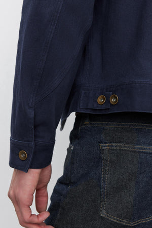 Close-up of a person wearing a Velvet by Graham & Spencer STEPHAN JACKET with metal buttons on the cuff and waist side adjusters, and dark blue jeans with visible stitching on the back pocket.