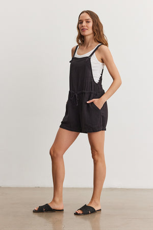 Woman in a casual black Velvet by Graham & Spencer Cyndee Cotton Gauze Romper and white tank top, standing in a minimalist space, wearing black slide sandals.