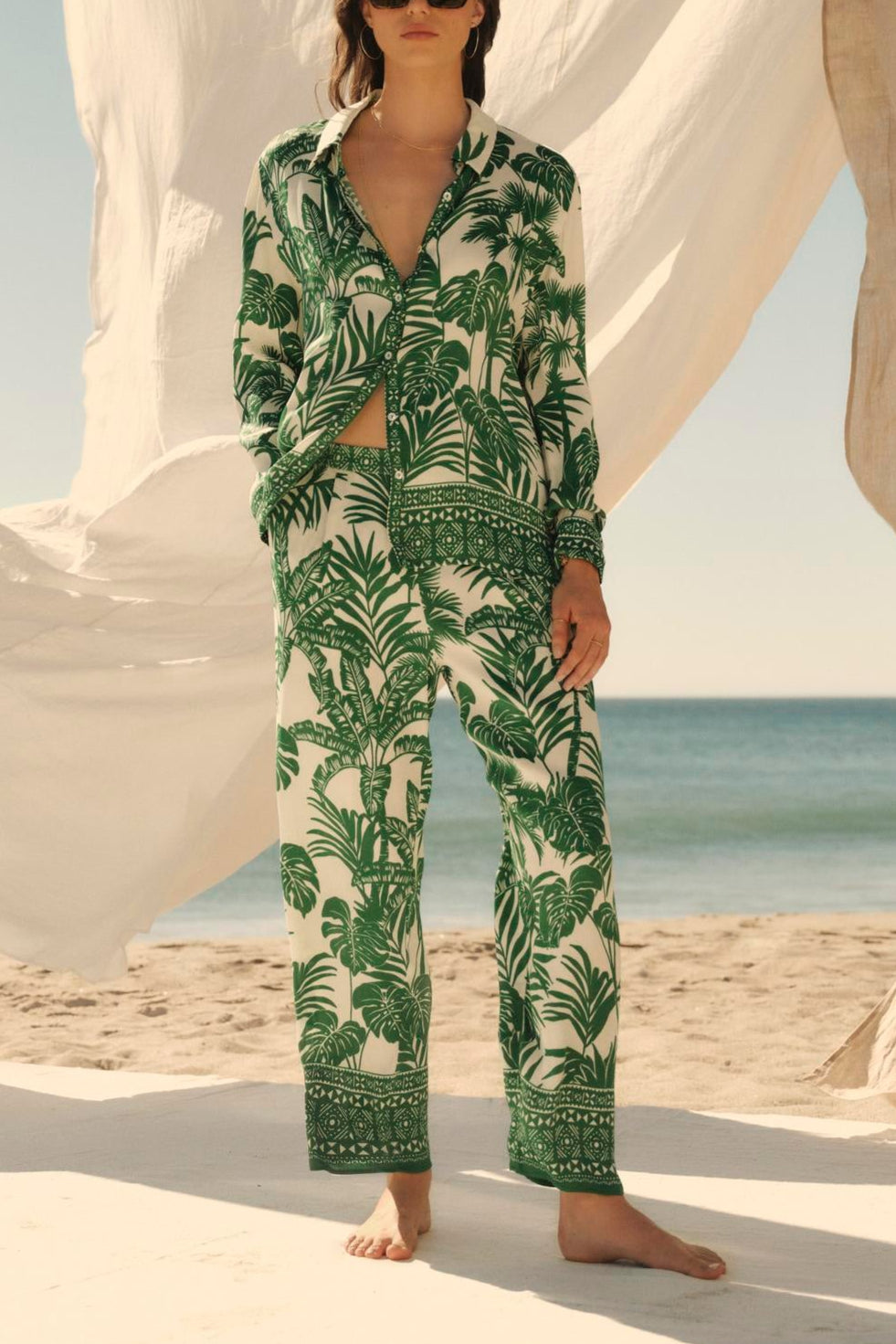 A woman in a green Velvet by Graham & Spencer AYLA button-up shirt stands on a beach, flanked by billowing white fabrics, with sunglasses on.