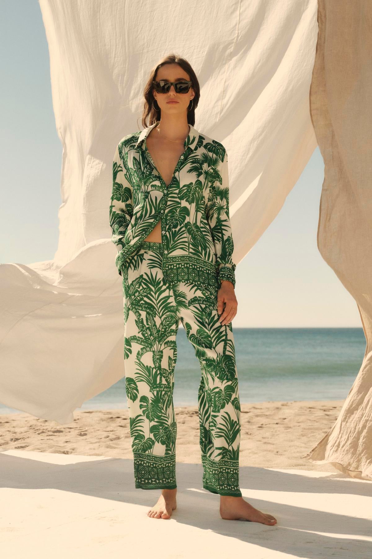 A woman in Velvet by Graham & Spencer's IRIS PANT with an elastic waist stands on a sandy beach, sunglasses on, with large white fabric sheets billowing in the breeze behind her.