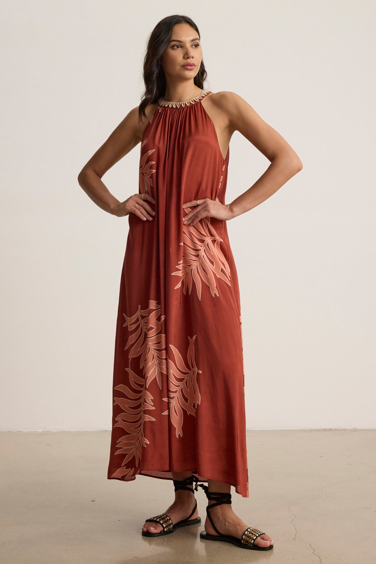 A woman stands indoors wearing the ODESSA DRESS by Velvet by Graham & Spencer, a sleeveless rust-colored maxi with a leaf print, paired with black flat sandals and her hair down.-39123393740993