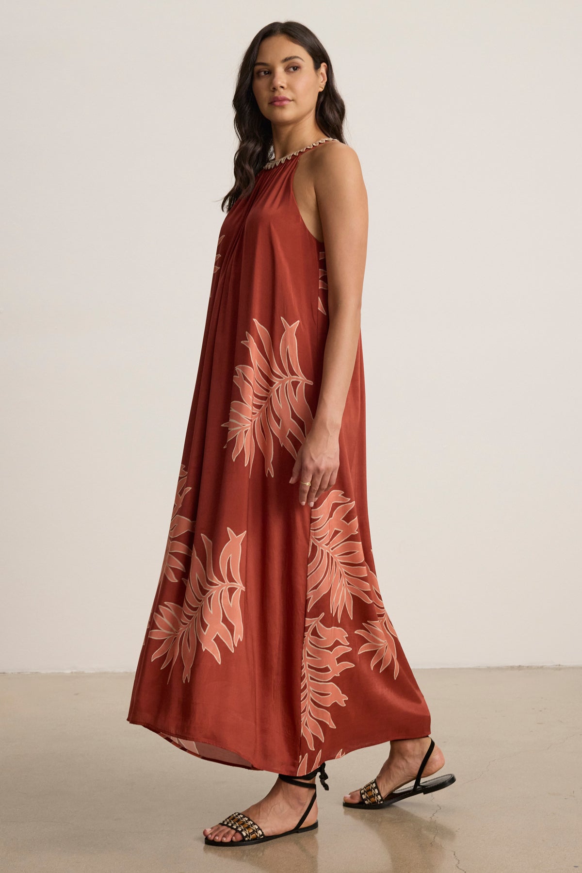 A woman stands indoors wearing the ODESSA DRESS by Velvet by Graham & Spencer—a sleeveless, rust maxi dress with beige leaf prints, subtle embroidery, and black flat sandals.-39123393708225