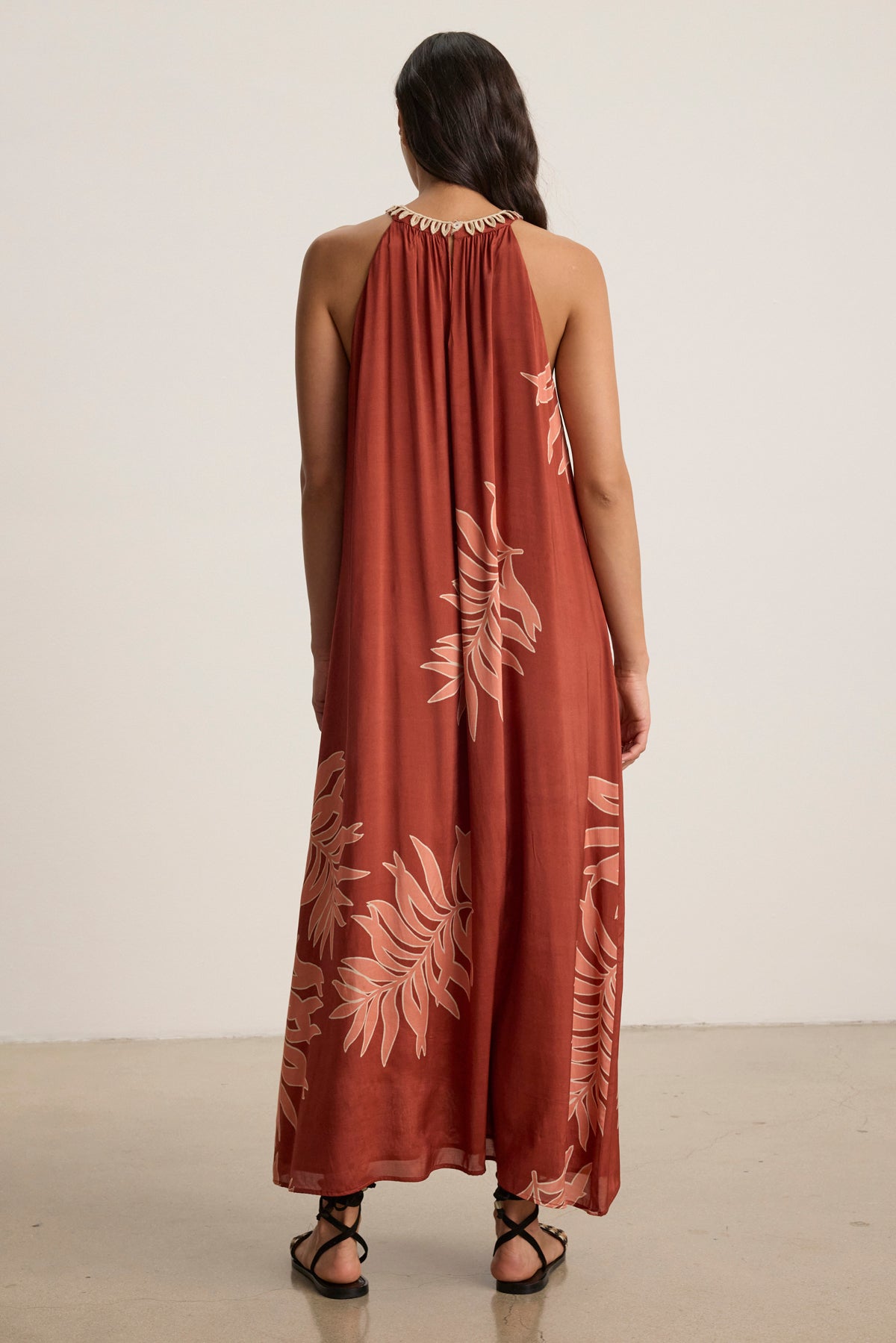 A woman with long dark hair, seen from behind on a plain background, wears the ODESSA DRESS by Velvet by Graham & Spencer—a sleeveless rust-colored maxi dress featuring a pale leaf print.-39123393675457