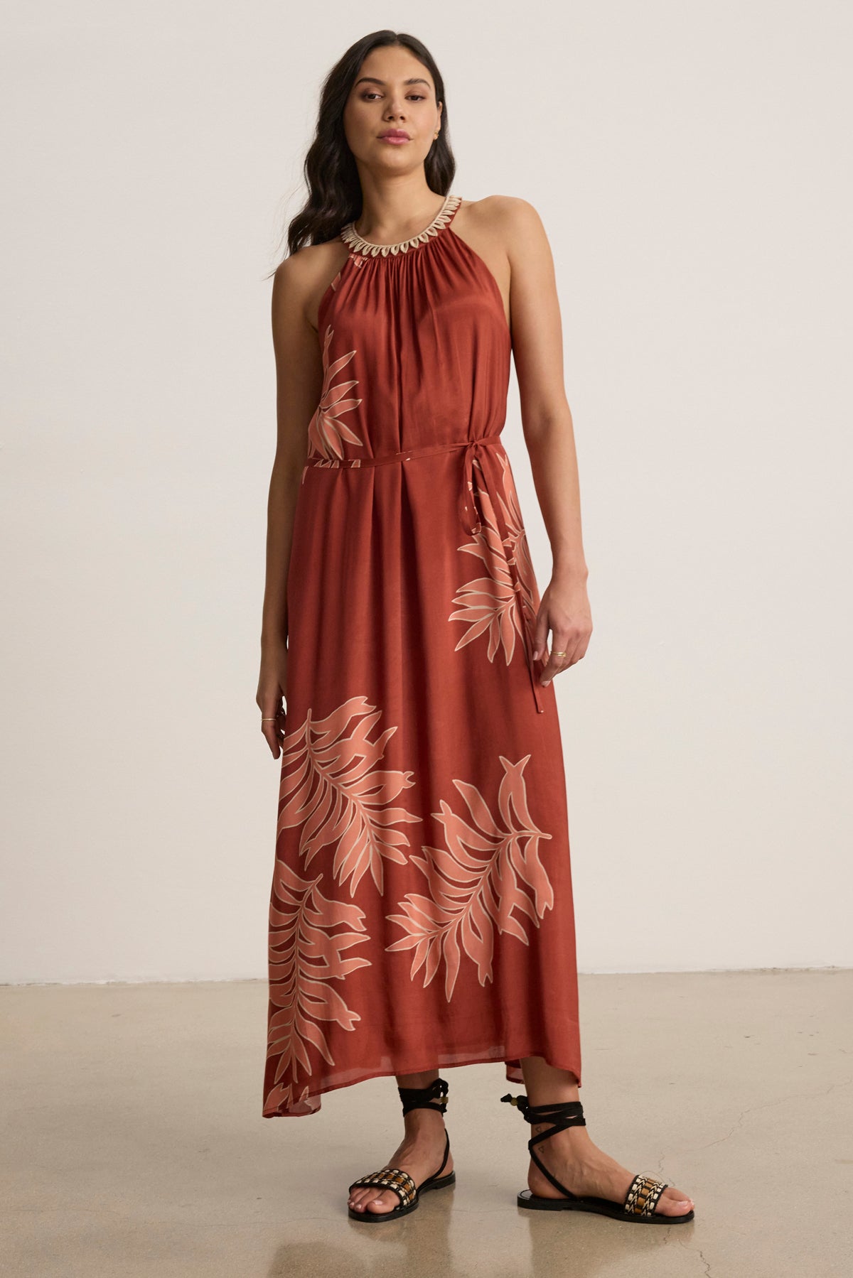 A woman faces the camera indoors against a plain background, wearing the ODESSA DRESS by Velvet by Graham & Spencer—a sleeveless rust maxi dress with leaf prints—paired with black sandals.-39123393642689
