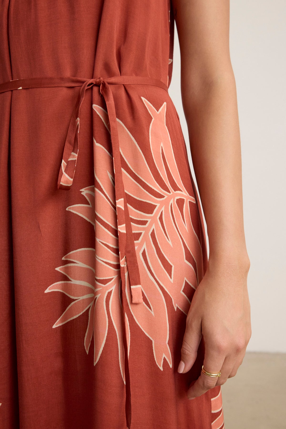 A close-up of a person wearing the ODESSA DRESS by Velvet by Graham & Spencer, a rust-colored floral maxi dress with a waist tie, showing their right hand adorned with two thin rings.-39123393609921