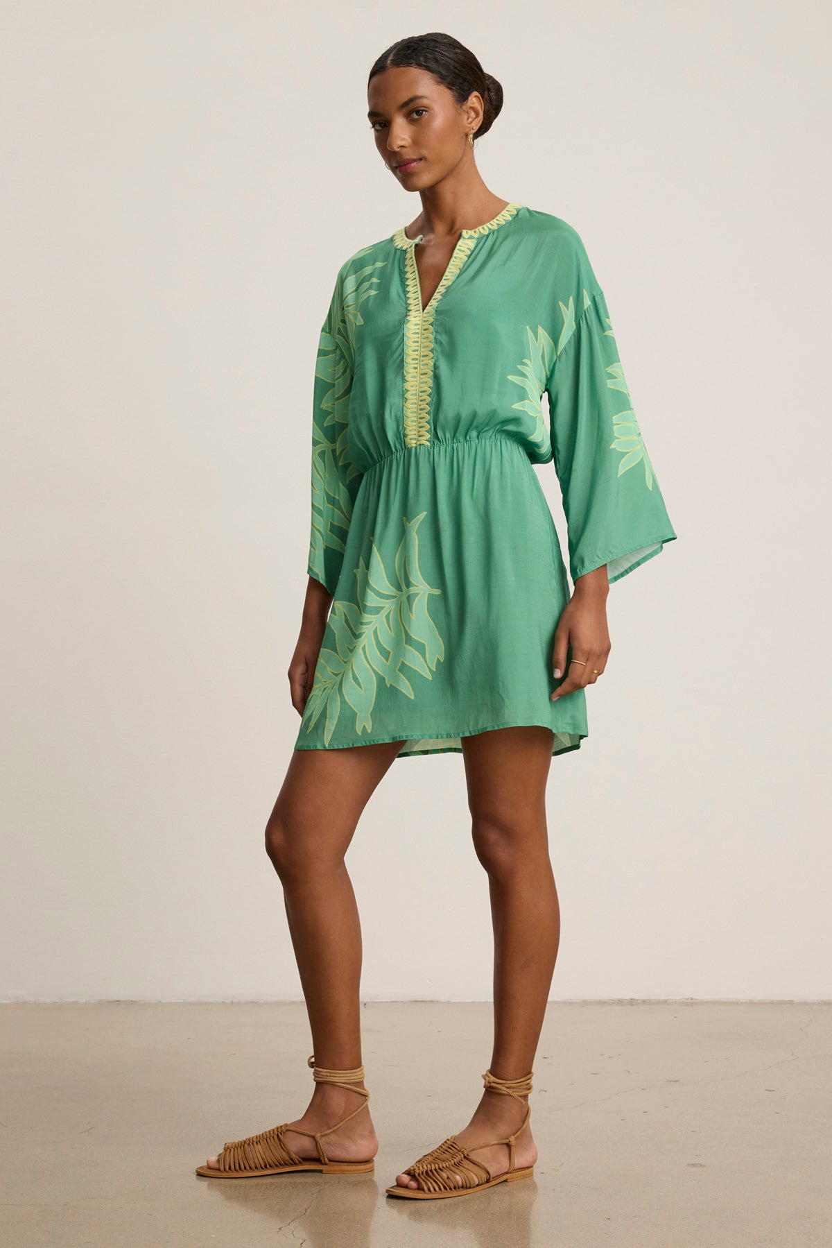 A woman stands indoors wearing the PHOEBE DRESS by Velvet by Graham & Spencer, a green long-sleeved V-neck mini dress with palmetto and leaf prints, paired with tan woven sandals.-39120133193921