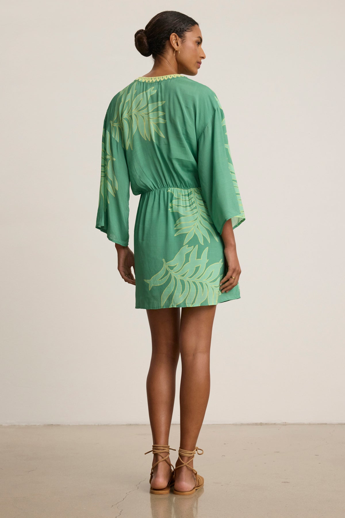 A woman faces away on a neutral background, wearing the Velvet by Graham & Spencer PHOEBE DRESS—a green mini dress with a yellow palmetto print—paired with tan sandals.-39120133161153