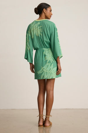 A woman faces away on a neutral background, wearing the Velvet by Graham & Spencer PHOEBE DRESS—a green mini dress with a yellow palmetto print—paired with tan sandals.