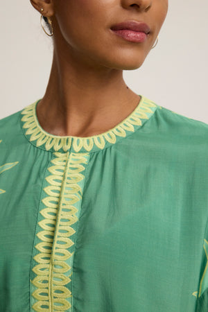 A person is seen wearing the PHOEBE DRESS by Velvet by Graham & Spencer, a green top with yellow embroidered details and an adjustable neckline; only the lower face and upper torso are shown.