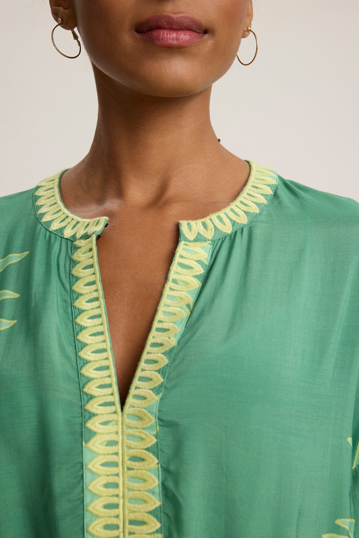 Close-up of a woman wearing the PHOEBE DRESS by Velvet by Graham & Spencer, a green mini dress with yellow embroidered trim on the adjustable neckline and front opening, styled with gold hoop earrings.-39120133095617