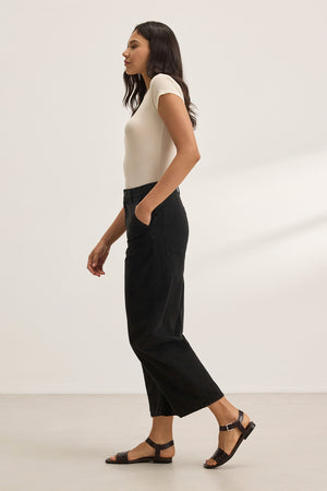 An individual wearing a white t-shirt and the ALEXIS PANT by Velvet by Graham & Spencer stands against a plain background, hands in pockets, and wears black sandals.