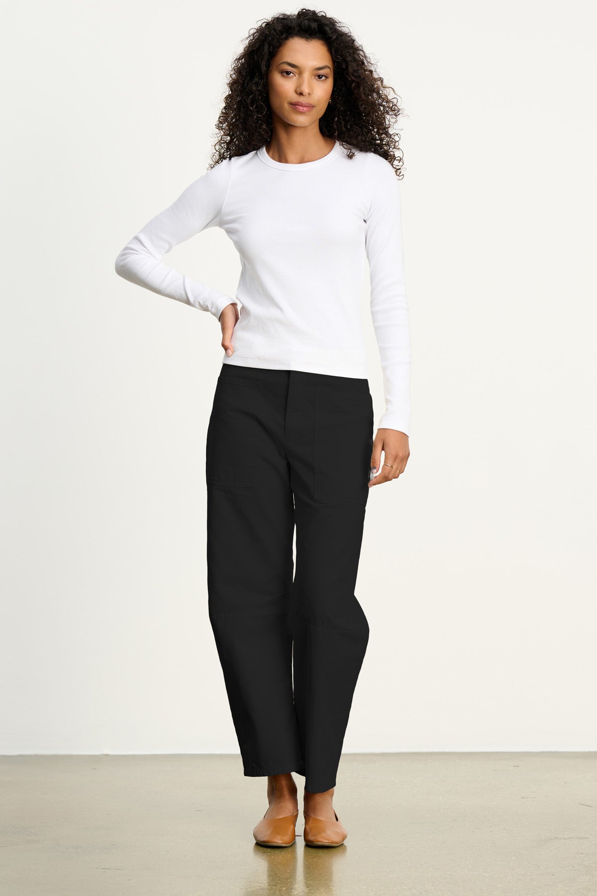 A woman stands against a plain background, wearing a long-sleeve white shirt, brown flat shoes, and the BRYLIE SANDED TWILL UTILITY PANT by Velvet by Graham & Spencer.-40435423740097