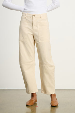 A person wears the BRYLIE SANDED TWILL UTILITY PANT by Velvet by Graham & Spencer, featuring cream cotton twill with patch pockets, paired with a white long-sleeve top and tan flats against a plain background.