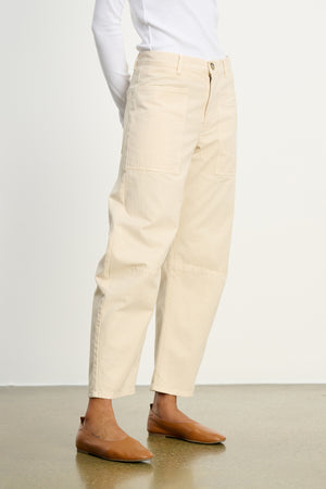 A person wears the BRYLIE SANDED TWILL UTILITY PANT by Velvet by Graham & Spencer, featuring off-white cotton twill with patch pockets, styled with a white long-sleeve top and brown flats against a plain background.