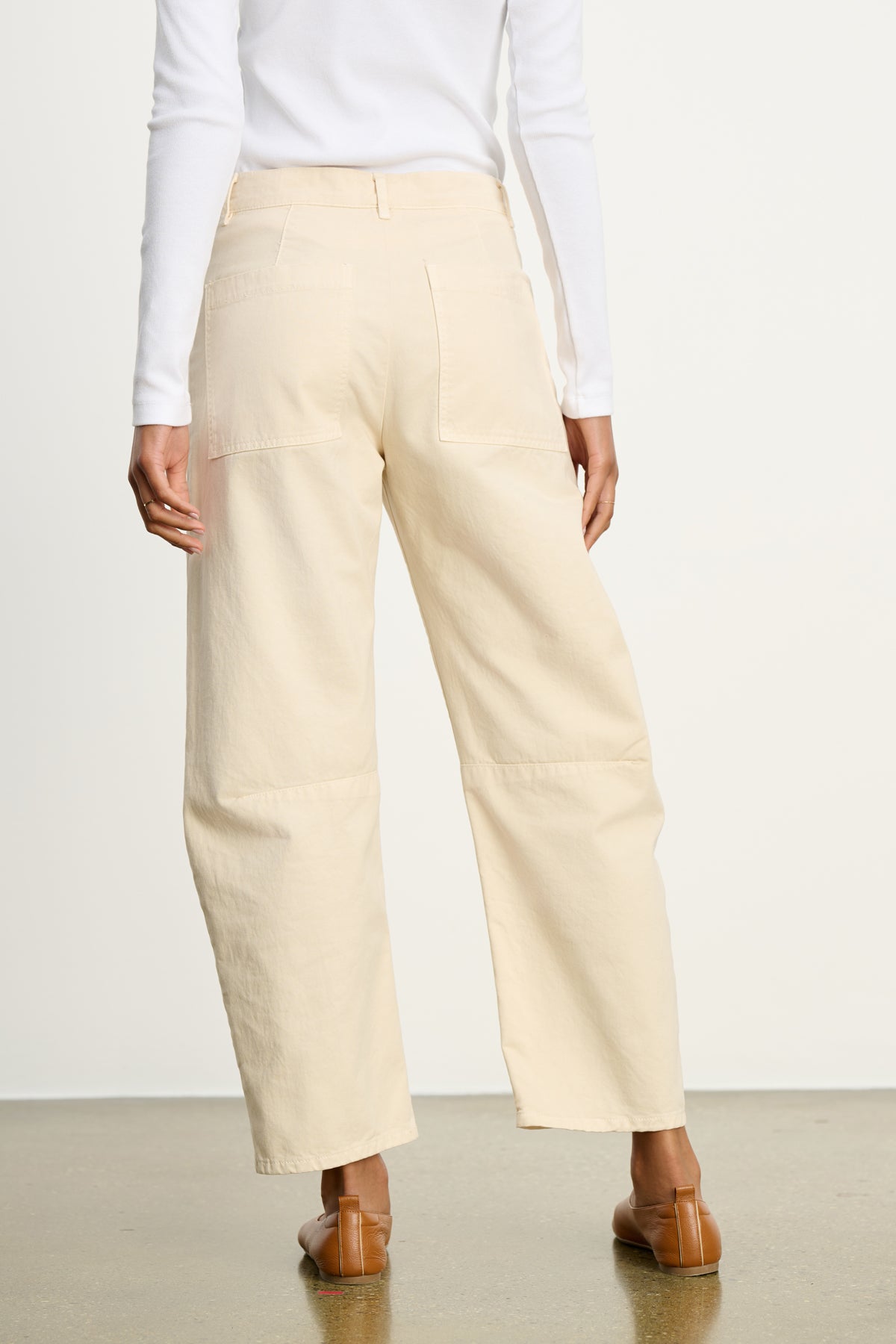 A person is shown from the back, wearing a white long-sleeve top and Velvet by Graham & Spencer's BRYLIE SANDED TWILL UTILITY PANT in beige, featuring patch pockets, paired with brown flat shoes on a polished floor.