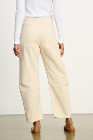 A person is shown from the back, wearing a white long-sleeve top and Velvet by Graham & Spencer's BRYLIE SANDED TWILL UTILITY PANT in beige, featuring patch pockets, paired with brown flat shoes on a polished floor.