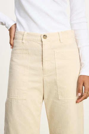 Model wears the BRYLIE SANDED TWILL UTILITY PANT by Velvet by Graham & Spencer—high-waisted beige cotton pants with patch pockets—paired with a white long-sleeve top for a modern, curved silhouette.