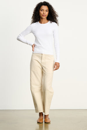 A woman stands against a plain background wearing a long-sleeve white shirt and the BRYLIE SANDED TWILL UTILITY PANT by Velvet by Graham & Spencer, featuring utilitarian patch pockets, paired with brown flats.