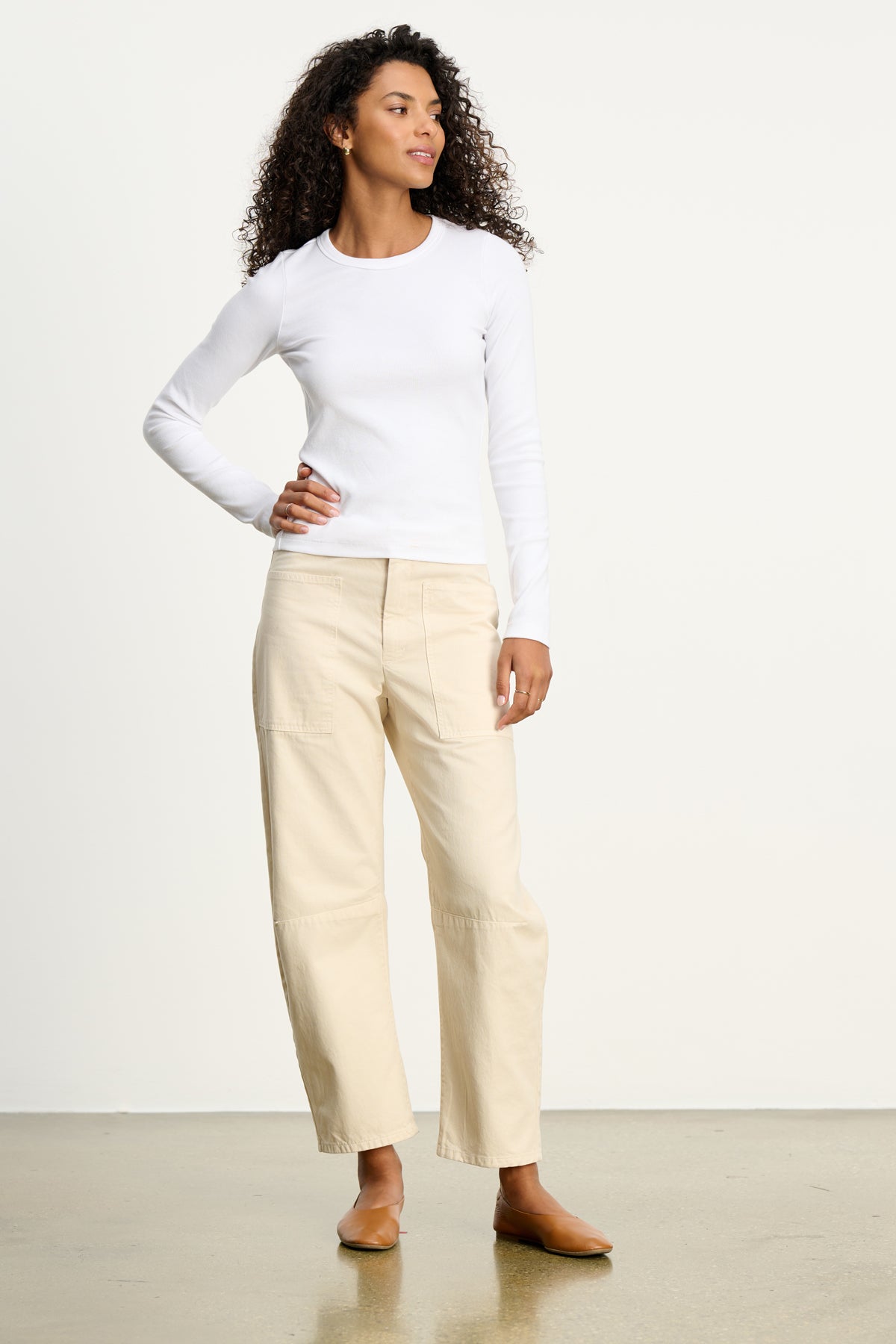 A woman stands indoors wearing a fitted white long-sleeve top and Velvet by Graham & Spencer’s BRYLIE SANDED TWILL UTILITY PANT, featuring seam details and patch pockets. She pairs the beige trousers with tan flats, her curly hair framing a softly curved silhouette.