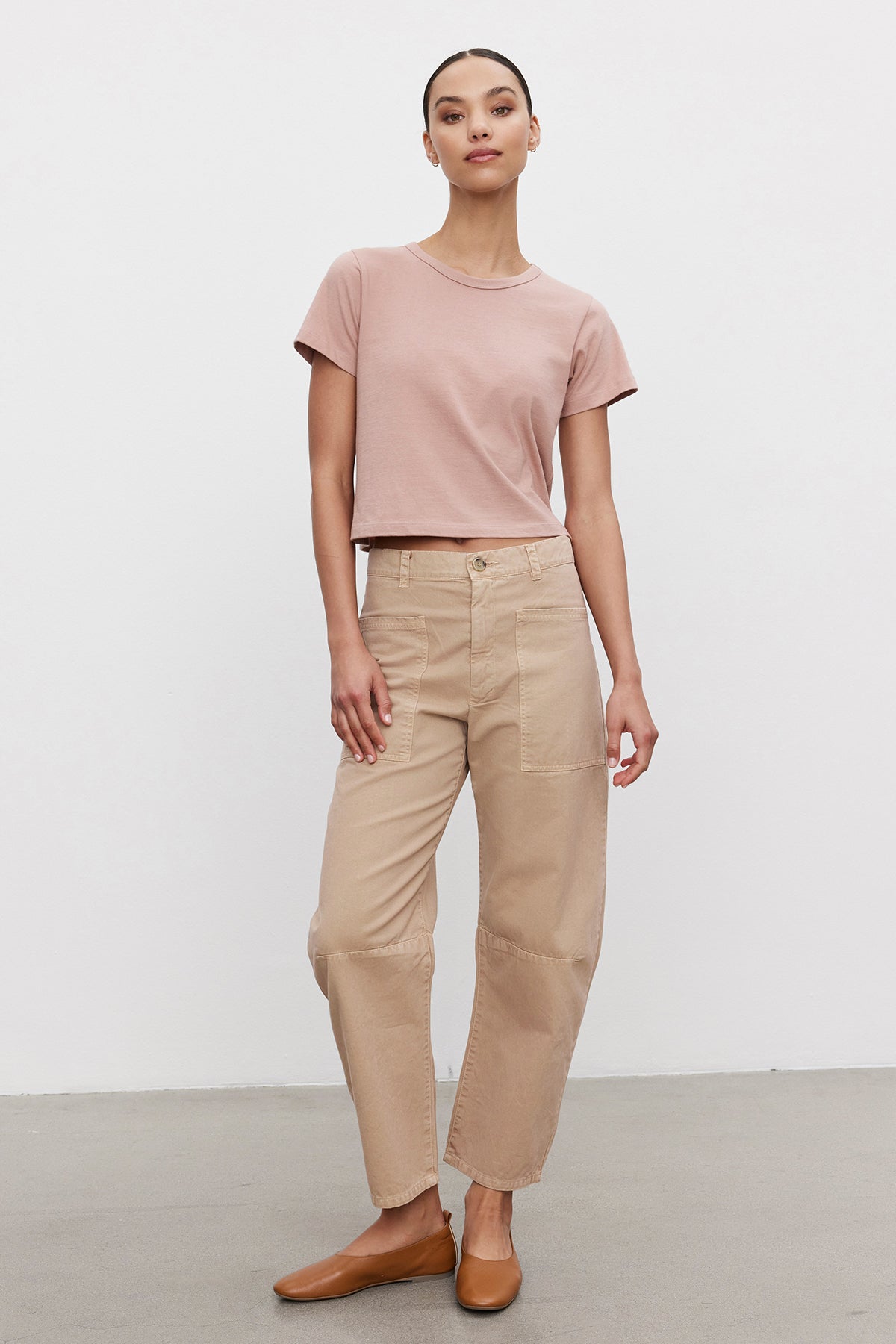 A woman stands against a plain background, wearing a light pink cropped t-shirt, Velvet by Graham & Spencer BRYLIE SANDED TWILL UTILITY PANT, and brown slip-on shoes.-39301429100737
