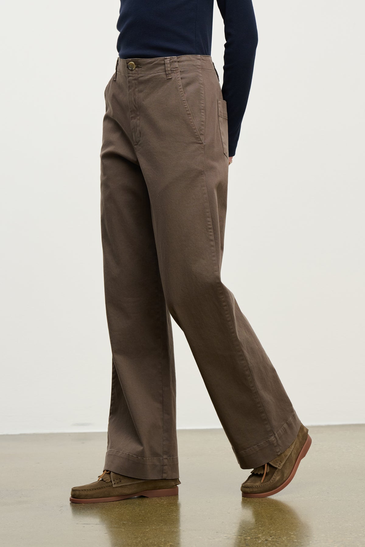 A person walks on a polished floor against a plain light wall, wearing Velvet by Graham & Spencer's DEN PANT—loose-fitting, wide-leg cotton twill pants with an elastic waistband—paired with brown shoes and a dark long-sleeve top.