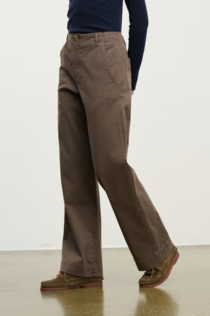 A person walks on a polished floor against a plain light wall, wearing Velvet by Graham & Spencer's DEN PANT—loose-fitting, wide-leg cotton twill pants with an elastic waistband—paired with brown shoes and a dark long-sleeve top.