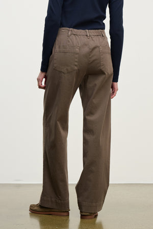 A person is shown from the back wearing the DEN PANT by Velvet by Graham & Spencer—wide-leg brown cotton twill pants with an elastic waistband—paired with a long-sleeve dark top, standing on a light floor against a plain background.
