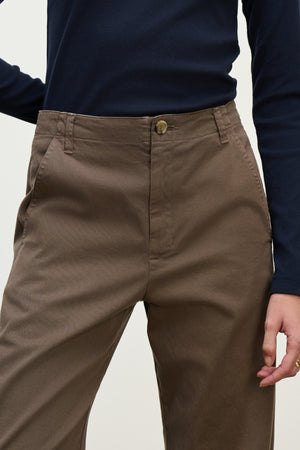 Model wears the DEN PANT by Velvet by Graham & Spencer in brown cotton twill, styled with a navy long-sleeve shirt. Shown from mid-torso to upper thighs, one hand is relaxed at their side.