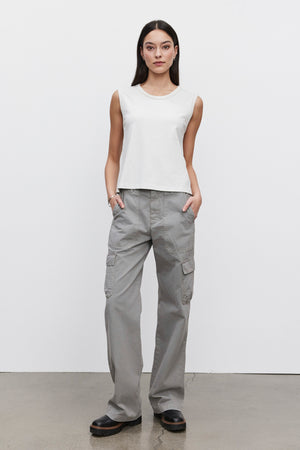 Against a plain white background, a woman stands wearing the MAKAYLA PANT from Velvet by Graham & Spencer—a pair of gray cotton twill cargo pants featuring practical cargo pockets. She pairs these with a white sleeveless shirt, showcasing an outfit that embodies both style and functionality in a versatile wardrobe.