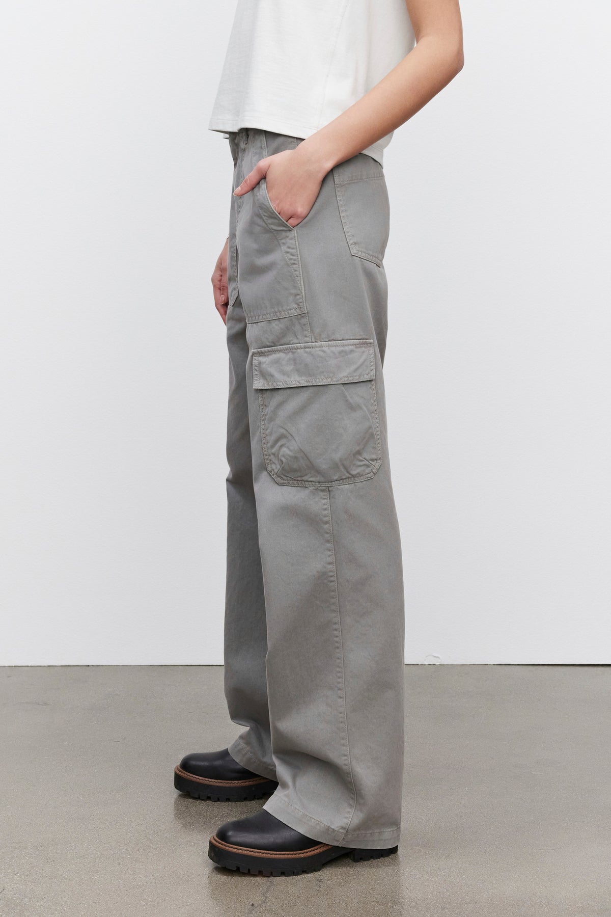 A person dressed in the MAKAYLA PANT by Velvet by Graham & Spencer, featuring gray cargo pants with flap pockets, paired with a white shirt. Brown shoes are showcased on a concrete floor, exemplifying a versatile wardrobe choice.-38372889264321