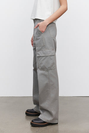 A person dressed in the MAKAYLA PANT by Velvet by Graham & Spencer, featuring gray cargo pants with flap pockets, paired with a white shirt. Brown shoes are showcased on a concrete floor, exemplifying a versatile wardrobe choice.