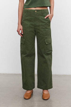 A person wearing Velvet by Graham & Spencer's MAKAYLA PANT, which are high-waisted and made from green sanded cotton twill, poses with hands in the front pockets. The pants feature two large cargo flap pockets and are paired with brown shoes, creating a versatile wardrobe choice that stands out against a plain white background.