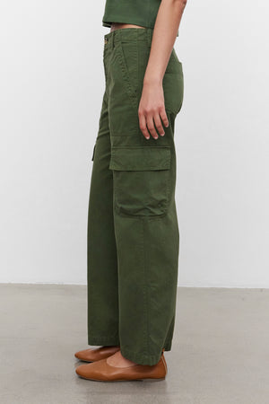 Someone is standing in a side profile view against a plain background wearing the MAKAYLA PANT by Velvet by Graham & Spencer, which features green cargo pants with flap pockets, and paired with brown shoes.