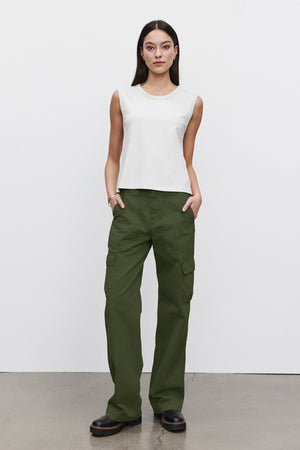 A woman stands against a plain white background, wearing a sleeveless white top, MAKAYLA PANT by Velvet by Graham & Spencer, and black shoes. Her hands are in her pockets.