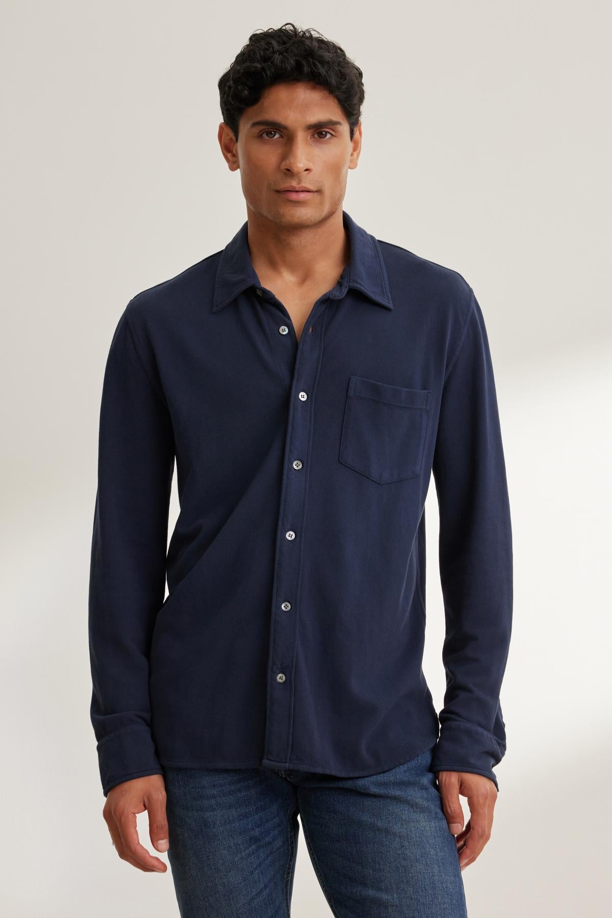 A person wearing a navy blue GIORGIO PIQUE BUTTON-UP SHIRT by Velvet by Graham & Spencer and blue jeans stands against a neutral background.