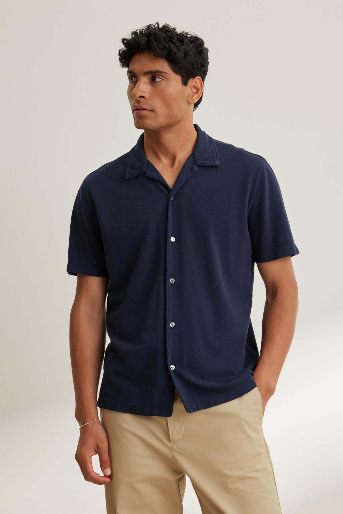 A man wearing a MARTYN PIQUE BUTTON-UP SHIRT by Velvet by Graham & Spencer pairs it with beige pants, glancing sideways against a plain backdrop.