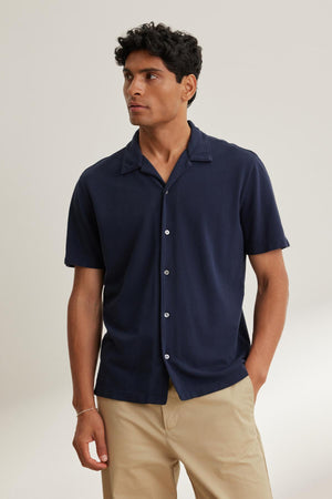 A man wearing a MARTYN PIQUE BUTTON-UP SHIRT by Velvet by Graham & Spencer pairs it with beige pants, glancing sideways against a plain backdrop.