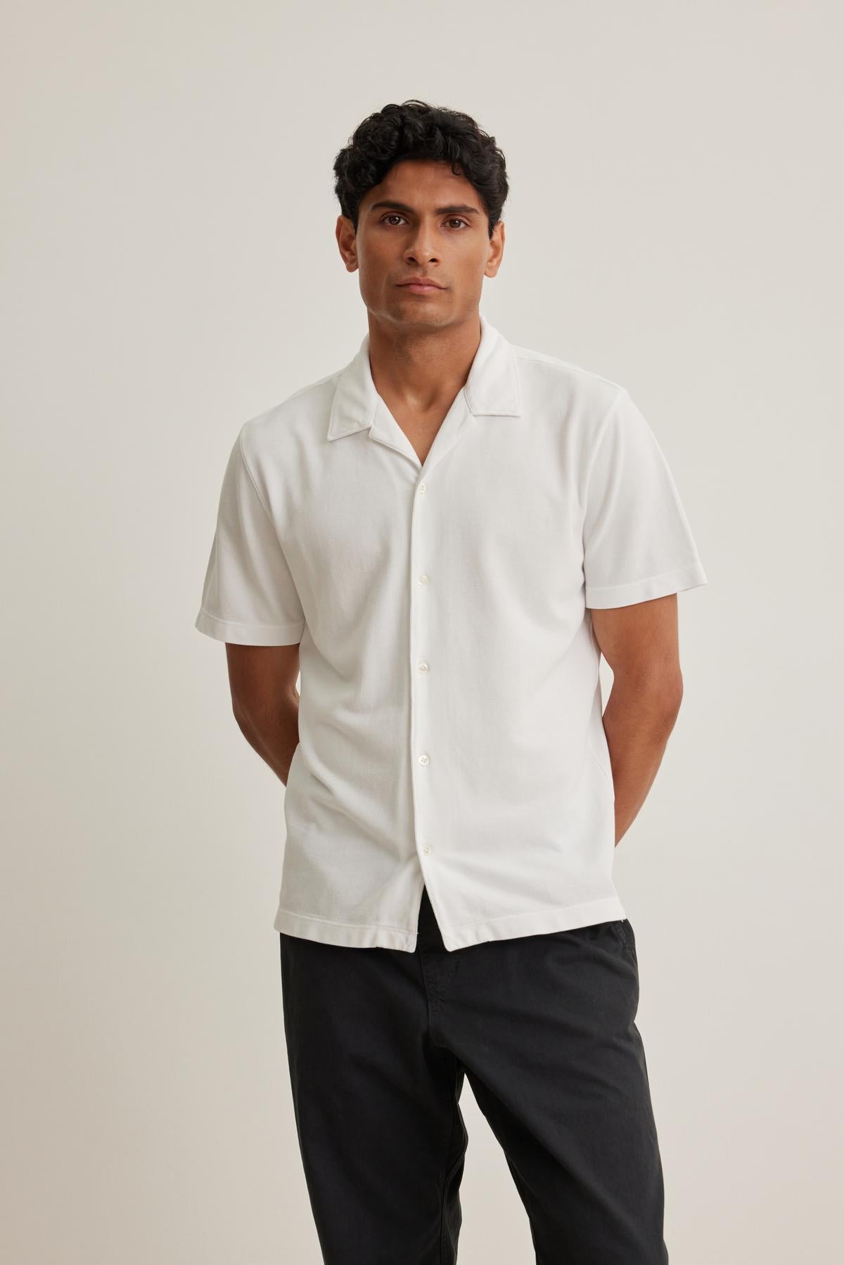Person standing against a plain backdrop, wearing the short-sleeve MARTYN PIQUE BUTTON-UP SHIRT by Velvet by Graham & Spencer and dark pants.