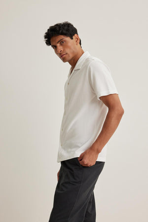 A man dressed in a MARTYN PIQUE BUTTON-UP SHIRT from Velvet by Graham & Spencer and dark pants stands against a plain background, gazing slightly to the side.