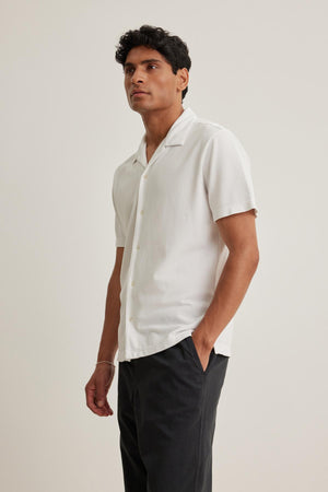 Someone wearing the MARTYN PIQUE BUTTON-UP SHIRT by Velvet by Graham & Spencer and dark pants stands with one hand in their pocket against a neutral background.