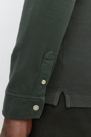 Close-up of a person wearing the MIKE POLO by Velvet by Graham & Spencer, a dark green piqué cotton long-sleeve shirt featuring textured fabric and buttoned cuffs, with visible side slits.
