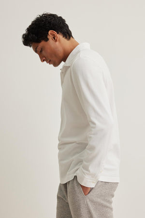 A person wearing the Velvet by Graham & Spencer SACHA POLO long-sleeve shirt and gray pants stands with hands in pockets, gazing down.